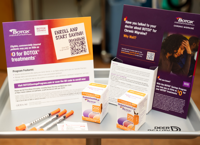 A metal tray holds promotional materials for Botox for Chronic Migraine, including two boxes of onabotulinumtoxinA, three syringes with orange caps, and several informational cards and brochures. One prominent card advertises that eligible, commercially insured patients may pay as little as $0 for Botox treatments and encourages enrollment and scanning a QR code to start saving.