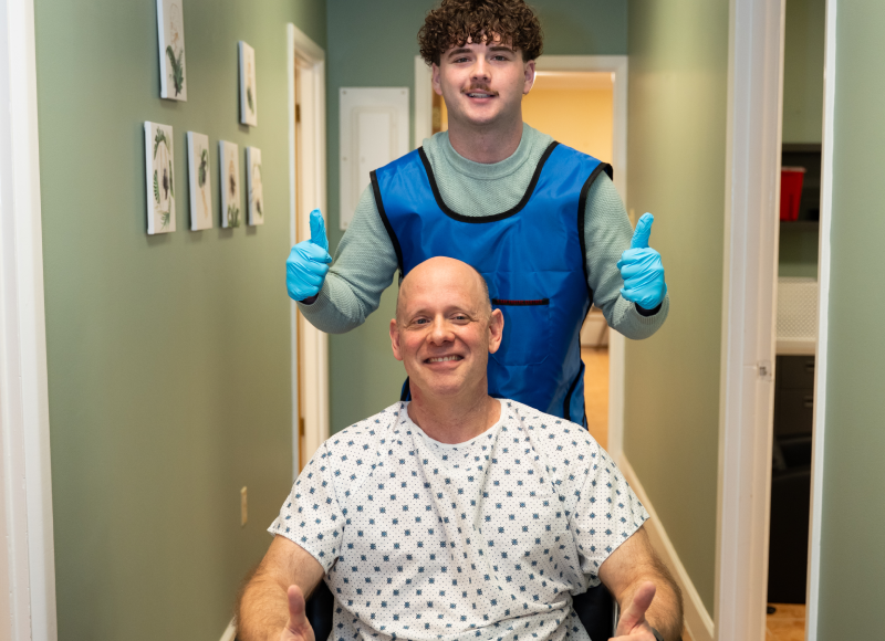 A young man with curly brown hair, wearing a blue protective vest and blue gloves, is pushing an older, fair-skinned man in a wheelchair down a hallway. Both men are smiling and giving thumbs-up gestures, suggesting a positive interaction or successful assistance.