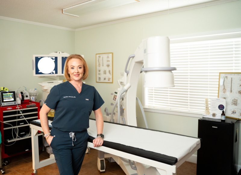 A female doctor in scrubs stands confidently next to a procedure table in a medical imaging room. The space features diagnostic equipment, anatomical charts, and a C-arm fluoroscopy machine used for spinal imaging.