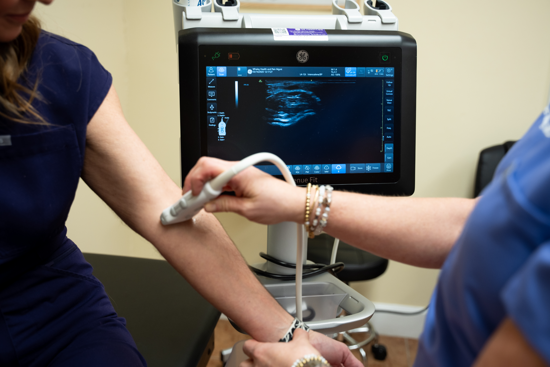 A medical professional uses an ultrasound device to examine a patient's forearm, with the ultrasound image visible on the monitor. The patient is seated on an exam table, and the procedure is taking place in a clinical setting.