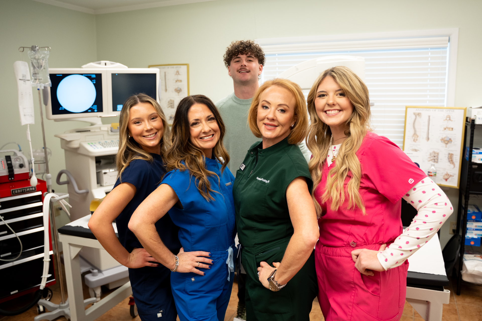 Five individuals, appearing to be healthcare professionals, are posing for a group photo in what seems to be a medical examination room with equipment in the background. The group consists of four women with fair skin and varying hair colors and a young man with fair skin and curly brown hair, all smiling towards the camera.