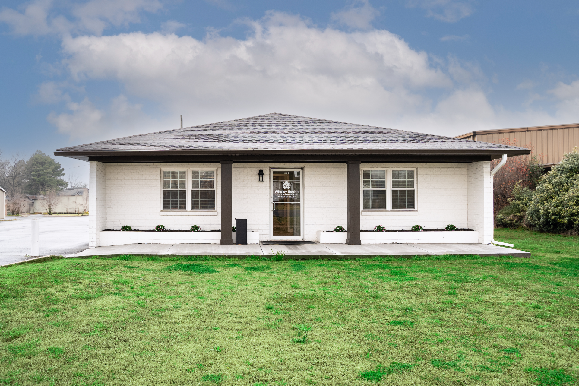 The image shows a white brick building with a dark gray roof and trim, featuring a glass entrance door with the words "Whaley Health & Pain Management" visible. The building is surrounded by a green lawn and small landscaping beds under a partly cloudy sky.
