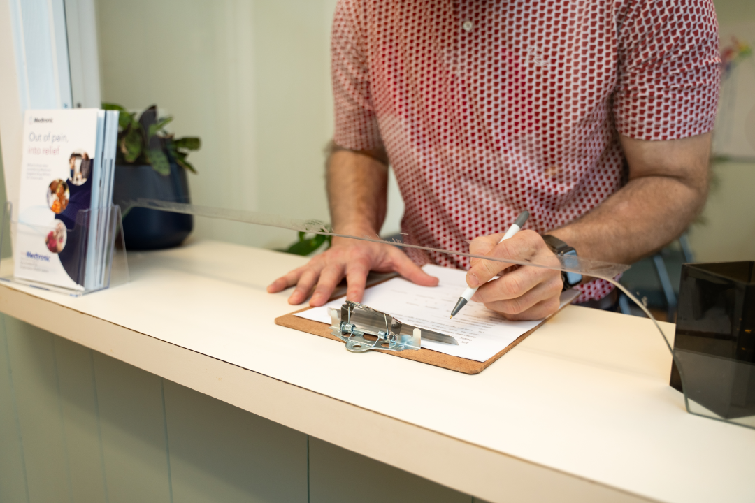A person with fair skin and visible arm hair is standing at a white counter, filling out a form on a clipboard with a white pen. They are wearing a red and white patterned short-sleeved shirt and a watch on their left wrist.