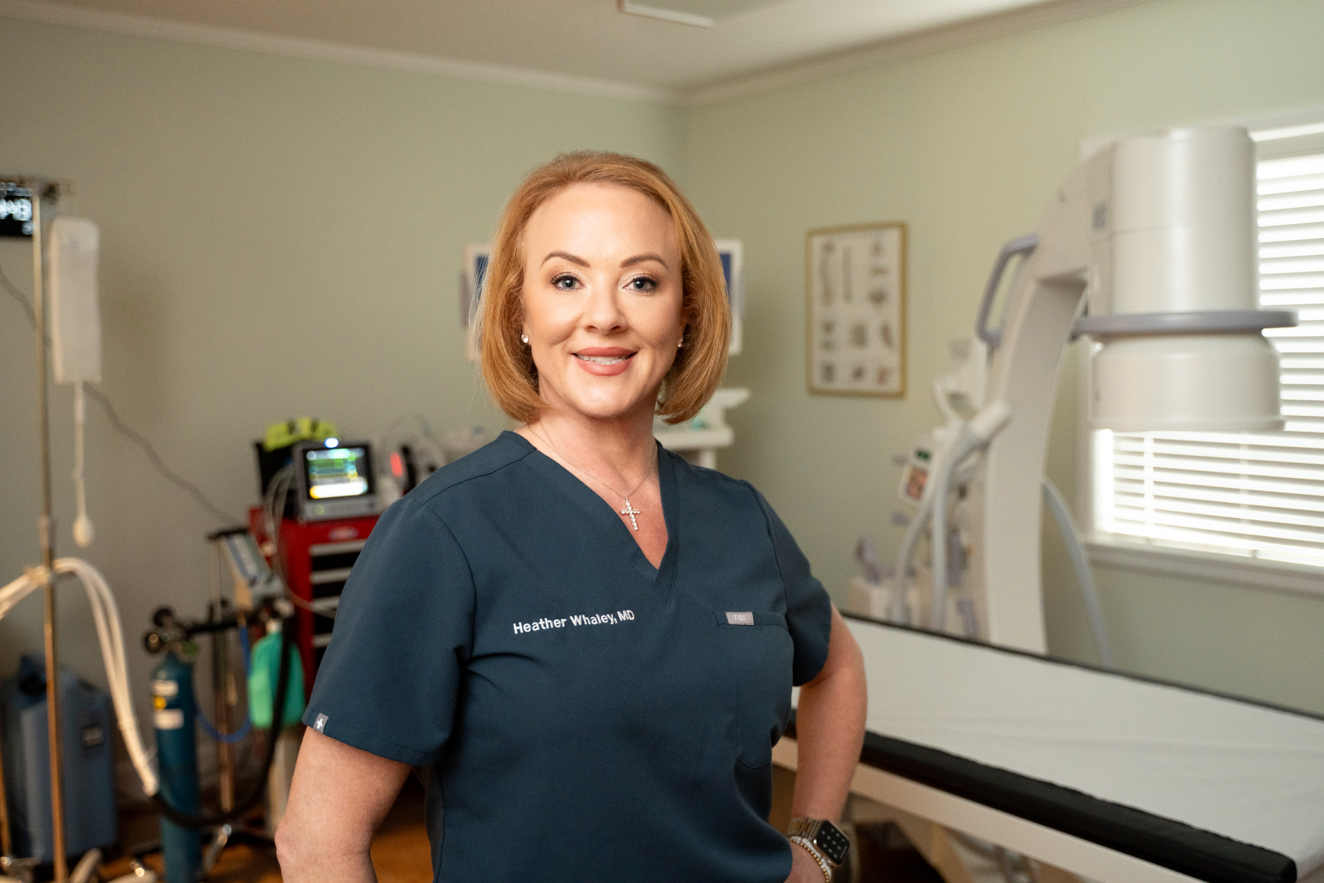 A fair-skinned woman with blonde hair, identified by the name "Heather Whaley, MD" embroidered on her teal scrub top, smiles at the camera. She appears to be in a medical or clinical setting, with medical equipment and a window visible in the background.