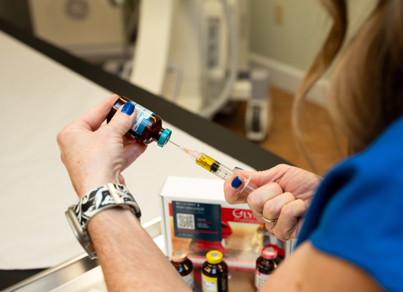 A person with fair skin and blue nail polish is shown from the shoulders down, preparing an injection. They are holding a syringe with a needle, drawing a yellow liquid from a small glass vial with a blue cap.