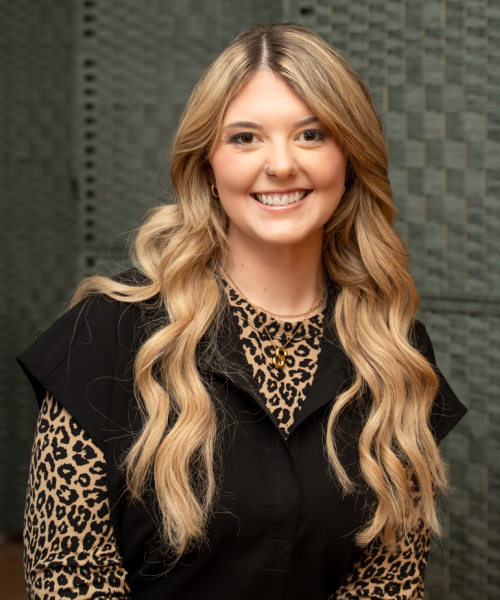 A smiling woman with long, wavy blonde hair and fair skin is looking directly at the camera. She is wearing a black sleeveless top over a long-sleeved shirt with a leopard print pattern.