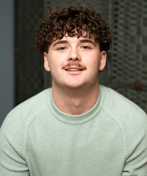 A young adult with fair skin, short curly brown hair, and a mustache is looking directly at the camera with a neutral expression. They are wearing a light green, textured crew-neck sweater.
