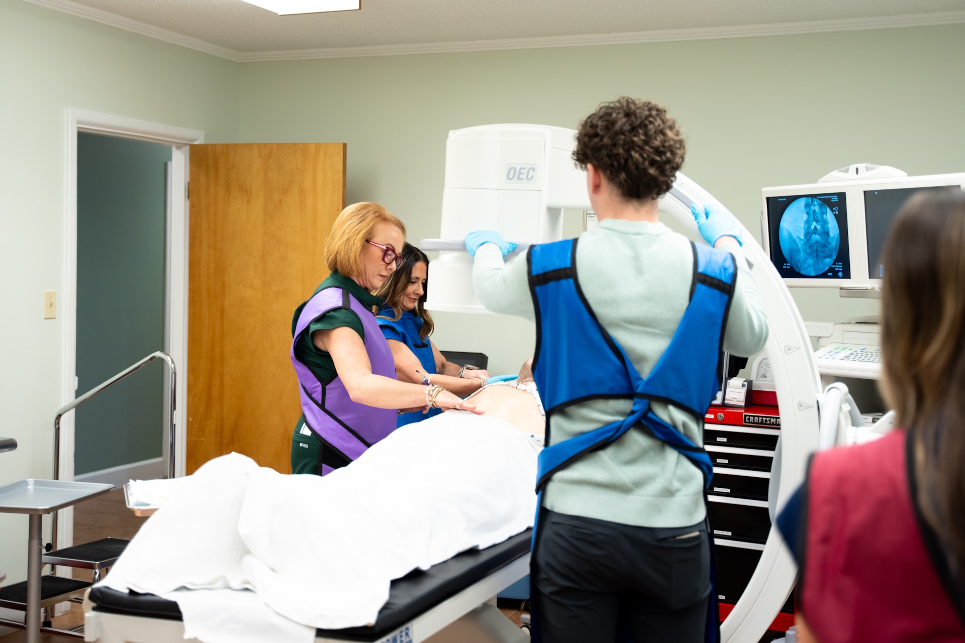 <br />
A patient lies face up on a medical table, covered by a white sheet, with a green blanket draped over their lower body. Their face is obscured by a large, dark gray or black shield, and a medical professional's hand is visible near the patient's head.