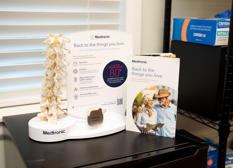 A pair of hands is shown drawing a yellow liquid from a small glass vial into a syringe. The person appears to be wearing a blue scrub top and a patterned watch or bracelet.