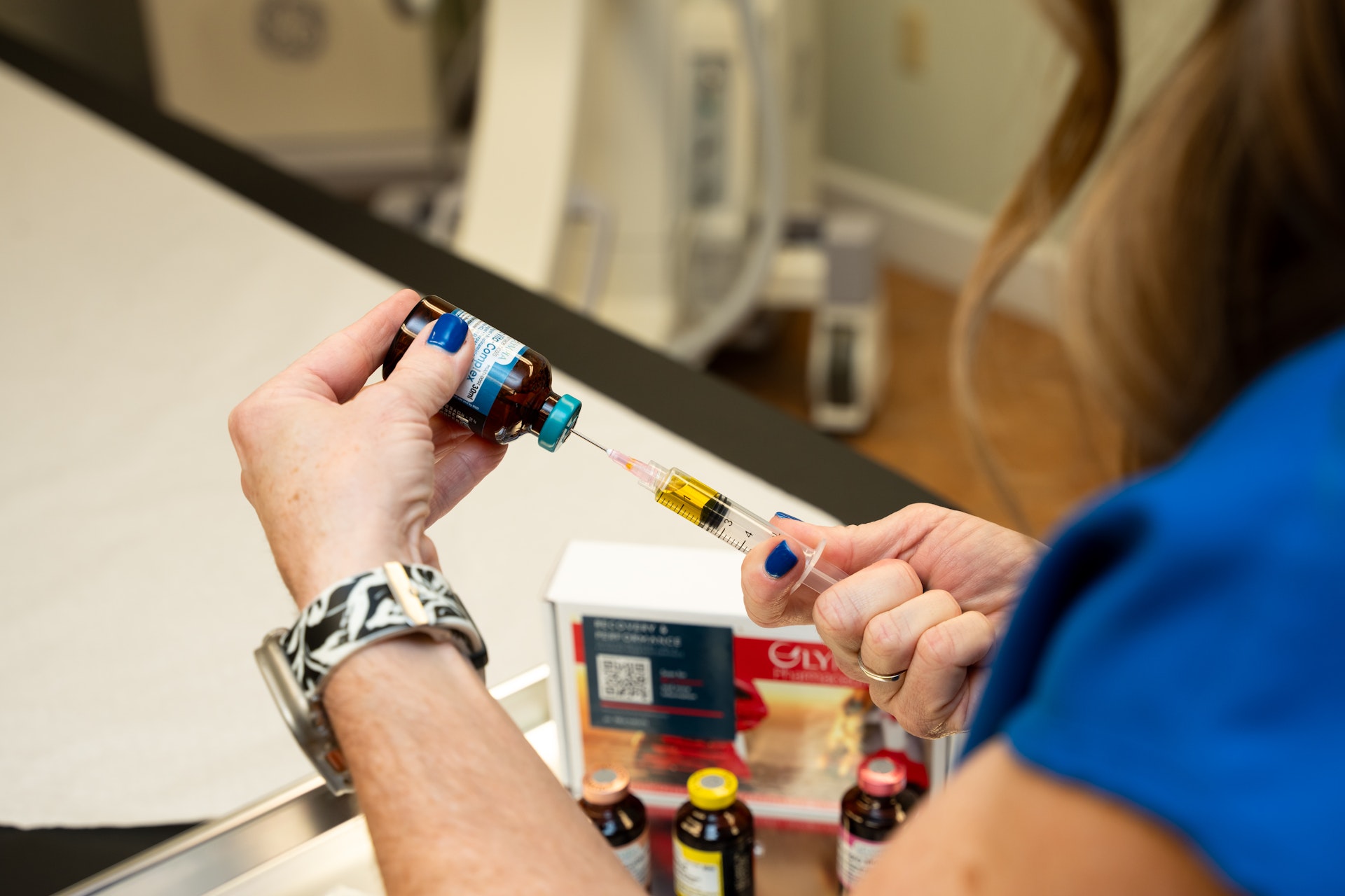 A medical professional uses an ultrasound device to examine a patient's forearm, with the ultrasound image visible on the monitor. The patient is seated on an exam table, and the procedure is taking place in a clinical setting.