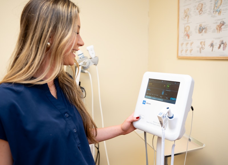 A young man with curly brown hair, wearing a blue protective vest and blue gloves, is pushing an older, fair-skinned man in a wheelchair down a hallway. Both men are smiling and giving thumbs-up gestures, suggesting a positive interaction or successful assistance.